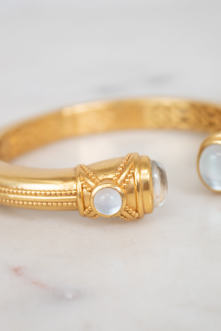 A gold bangle bracelet with ornate detailing and round white moonstone accents, resting on a light marble surface.