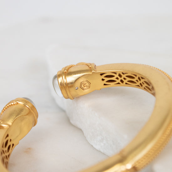 A gold-toned open cuff bangle bracelet with intricate interior filigree cutouts and a small moonstone accent, displayed on a white marble slab against a light background.