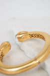 A gold-toned open cuff bangle bracelet with intricate interior filigree cutouts and a small moonstone accent, displayed on a white marble slab against a light background.
