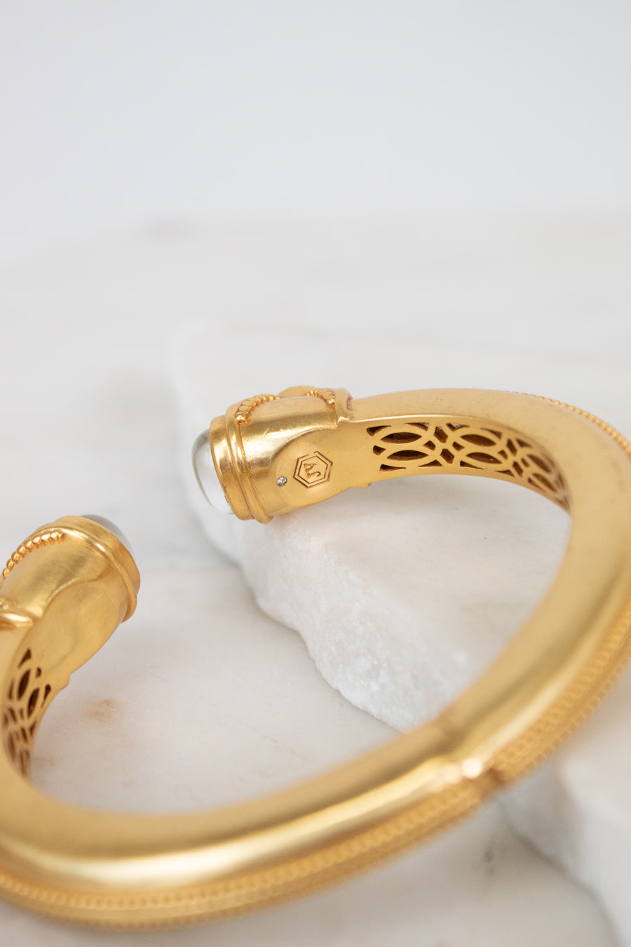 A gold-toned open cuff bangle bracelet with intricate interior filigree cutouts and a small moonstone accent, displayed on a white marble slab against a light background.