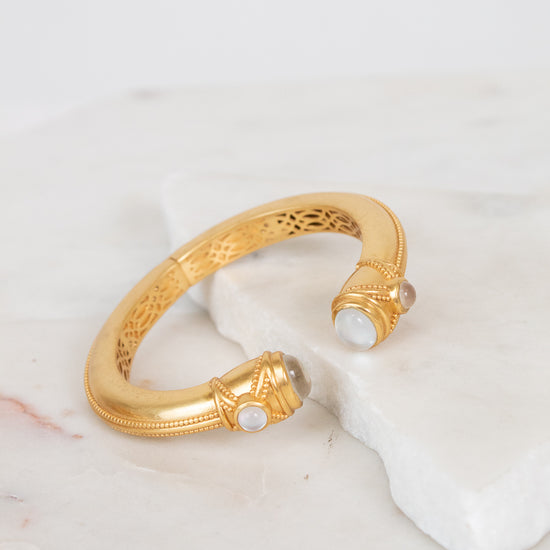 A gold-toned open cuff bracelet with intricate interior filigree detailing and small moonstone gemstone accents at the ends, displayed on a white marble slab against a light background.