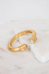 A gold-toned open cuff bracelet with intricate interior filigree detailing and small moonstone gemstone accents at the ends, displayed on a white marble slab against a light background.