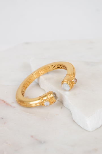 A gold-toned open cuff bracelet with intricate interior filigree detailing and small moonstone gemstone accents at the ends, displayed on a white marble slab against a light background.