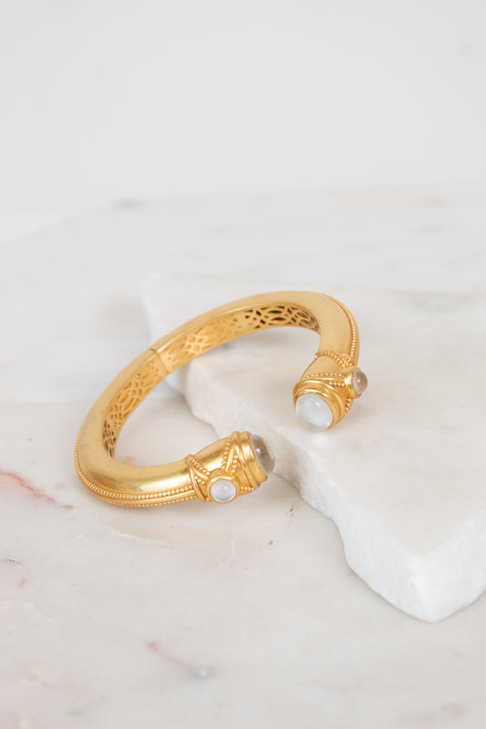 A gold-toned open cuff bracelet with intricate interior filigree detailing and small moonstone gemstone accents at the ends, displayed on a white marble slab against a light background.