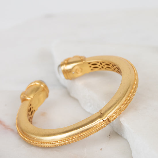A gold-toned open cuff bracelet with a textured outer edge and openwork filigree detailing inside, resting on a white marble slab against a white background.