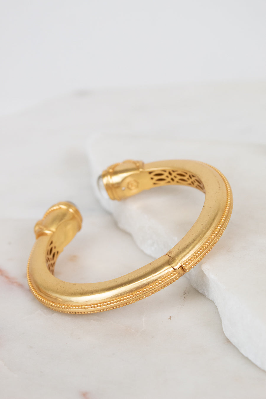 A gold-toned open cuff bracelet with a textured outer edge and openwork filigree detailing inside, resting on a white marble slab against a white background.