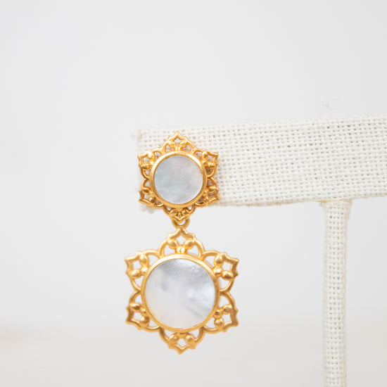 A pair of gold-toned statement earrings with oval white mother-of-pearl inlays, displayed on a white woven jewelry stand against a white background.