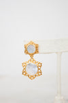 A pair of gold-toned statement earrings with oval white mother-of-pearl inlays, displayed on a white woven jewelry stand against a white background.