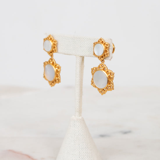 A pair of gold-toned statement earrings with oval white inlays, displayed on a white fabric jewelry stand against a light marble surface and white background.
