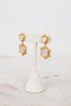 A pair of gold-toned statement earrings with oval white inlays, displayed on a white fabric jewelry stand against a light marble surface and white background.