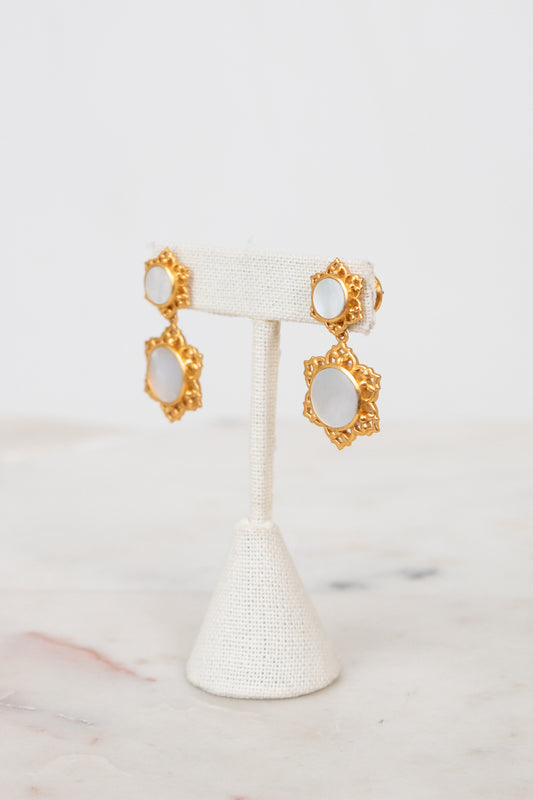 A pair of gold-toned statement earrings with oval white inlays, displayed on a white fabric jewelry stand against a light marble surface and white background.