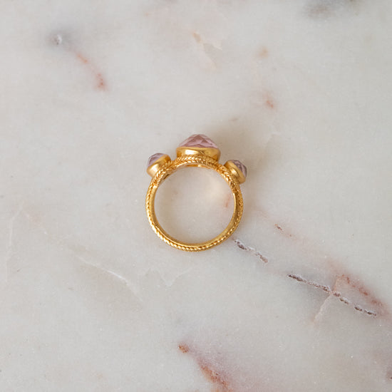 A gold ring with a textured band and a central pink glass stones, resting on a white marble surface.