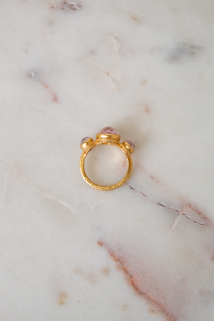 A gold ring with a textured band and a central pink glass stones, resting on a white marble surface.