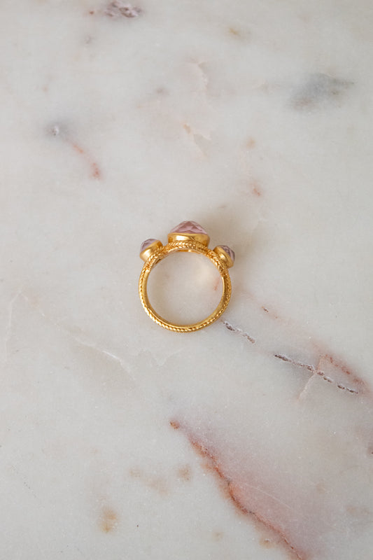 A gold ring with a textured band and a central pink glass stones, resting on a white marble surface.