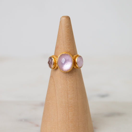 A gold ring with three oval light pink glass stones displayed on a light wood cone stand against a soft white background.