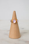 A gold ring with three oval light pink glass stones displayed on a light wood cone stand against a soft white background.