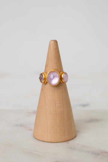 A gold ring with three oval light pink glass stones displayed on a light wood cone stand against a soft white background.