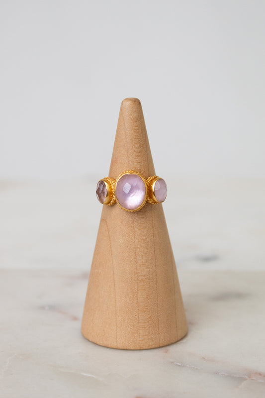 A gold ring with three oval light pink glass stones displayed on a light wood cone stand against a soft white background.