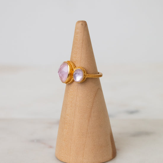 A gold ring with oval light pink glass stones displayed on a light wood cone stand against a soft white background.