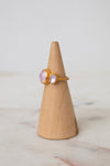A gold ring with oval light pink glass stones displayed on a light wood cone stand against a soft white background.
