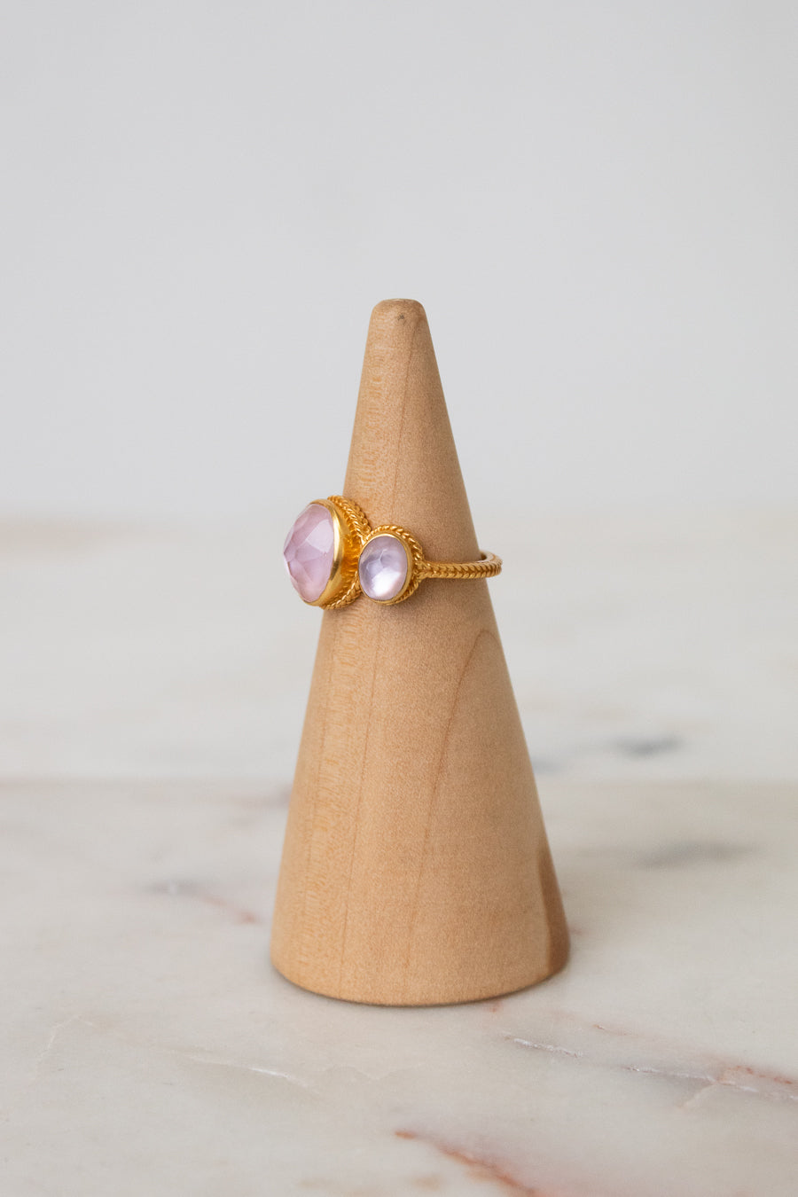 A gold ring with oval light pink glass stones displayed on a light wood cone stand against a soft white background.