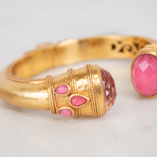A gold-toned bangle bracelet with pink glass stone accents, shown in a close-up shot on a light marble surface.