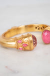 A gold-toned bangle bracelet with pink glass stone accents, shown in a close-up shot on a light marble surface.