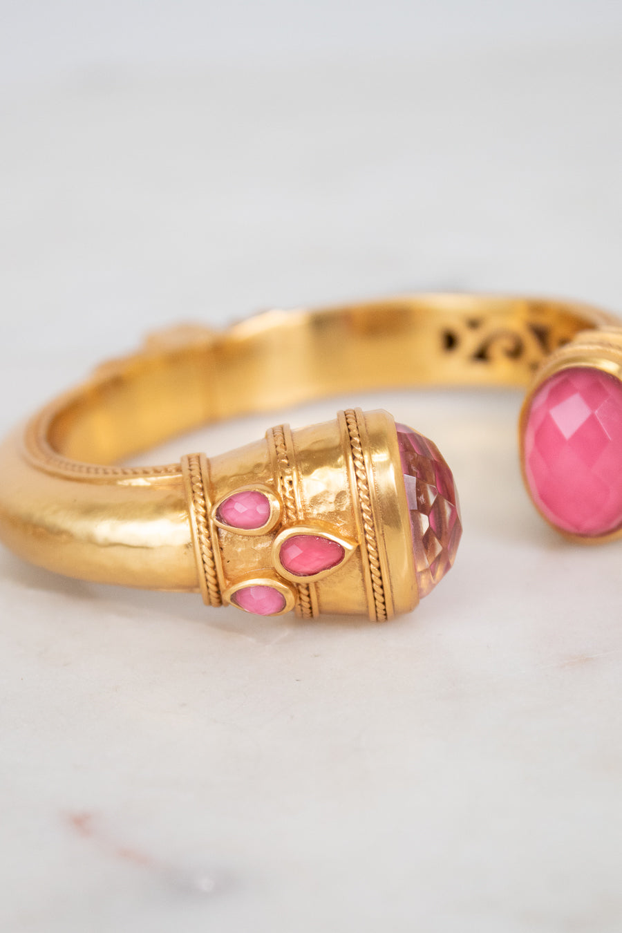 A gold-toned bangle bracelet with pink glass stone accents, shown in a close-up shot on a light marble surface.