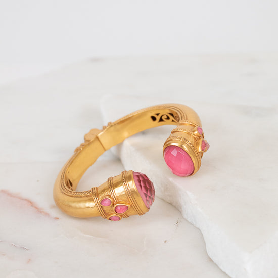 A gold-toned cuff bracelet with pink glass stone accents, displayed on a white marble slab against a light background.
