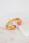 A gold-toned cuff bracelet with pink glass stone accents, displayed on a white marble slab against a light background.