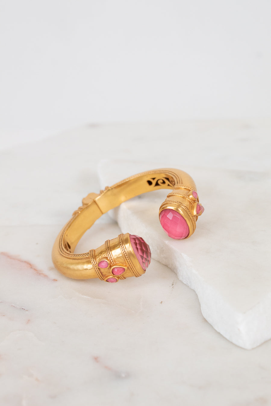 A gold-toned cuff bracelet with pink glass stone accents, displayed on a white marble slab against a light background.