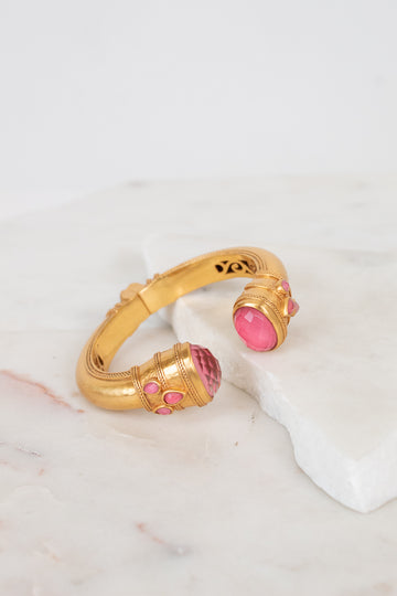 A gold-toned cuff bracelet with pink glass stone accents, displayed on a white marble slab against a light background.
