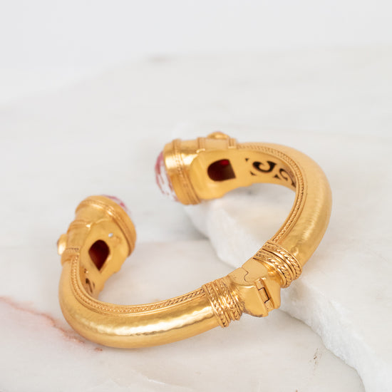 A gold-toned open cuff bracelet with decorative textured bands and small pink glass stone accents at the ends, displayed on a white marble surface.