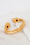 A gold-toned open cuff bracelet with decorative textured bands and small pink glass stone accents at the ends, displayed on a white marble surface.