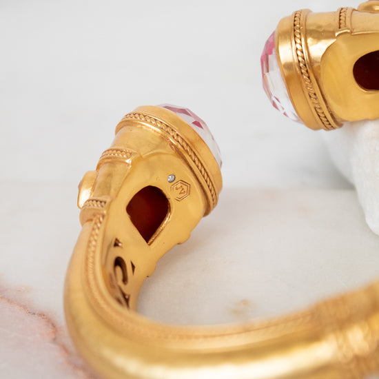 Gold-plated cuff bracelet with carved details and pinkish-red glass stones on the ends, displayed on a white marble surface.