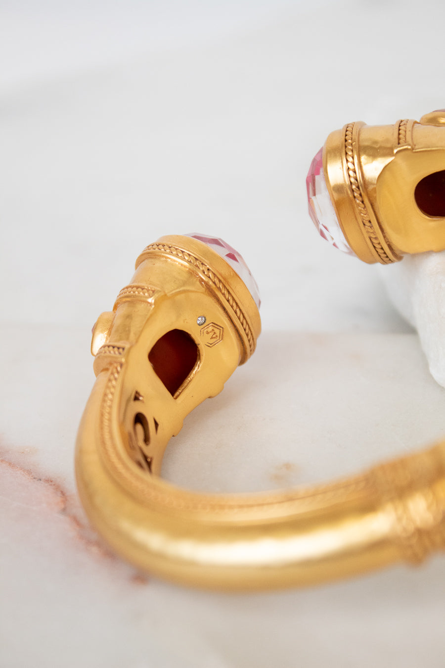 Gold-plated cuff bracelet with carved details and pinkish-red glass stones on the ends, displayed on a white marble surface.