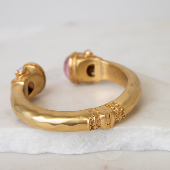 A gold-toned open cuff bracelet with decorative textured details and a pink gemstone accent, displayed on a white marble slab against a light gray background.