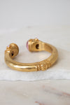A gold-toned open cuff bracelet with decorative textured details and a pink gemstone accent, displayed on a white marble slab against a light gray background.