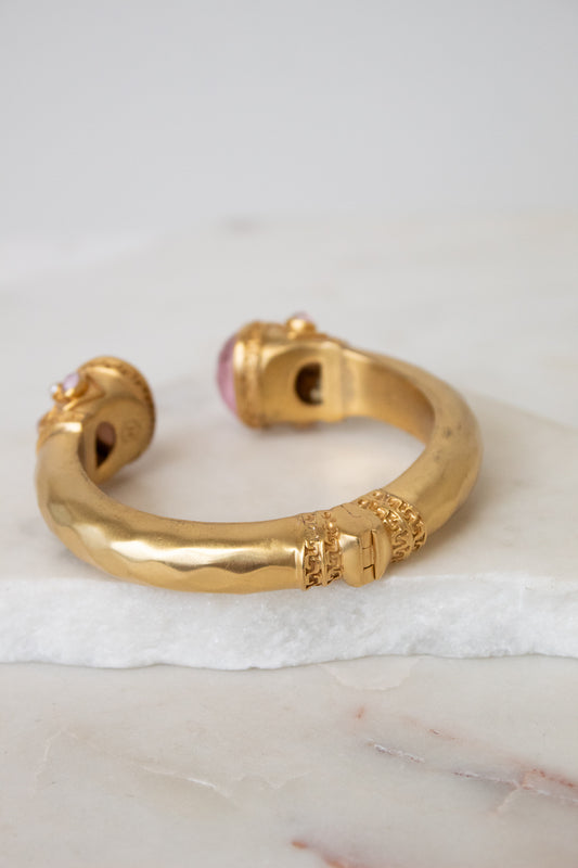 A gold-toned open cuff bracelet with decorative textured details and a pink gemstone accent, displayed on a white marble slab against a light gray background.