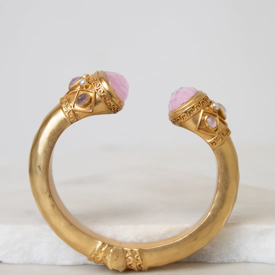 A gold-toned open cuff bracelet featuring two pink cabochon gemstones and small clear stones, displayed on a white marble surface against a light gray background.