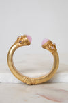 A gold-toned open cuff bracelet featuring two pink cabochon gemstones and small clear stones, displayed on a white marble surface against a light gray background.