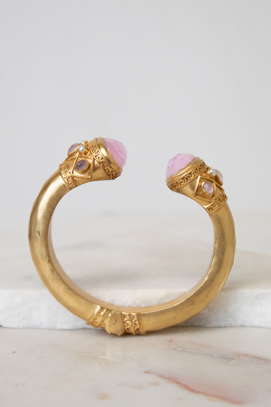A gold-toned open cuff bracelet featuring two pink cabochon gemstones and small clear stones, displayed on a white marble surface against a light gray background.