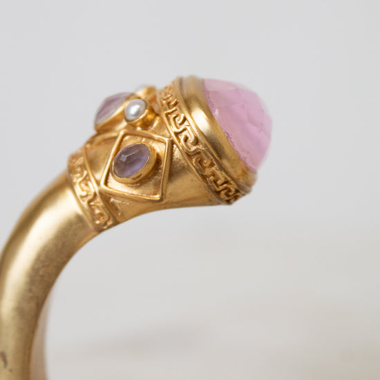 A close-up of a gold bracelet featuring a large pink cabochon stone and smaller purple and clear stones in an ornate setting.
