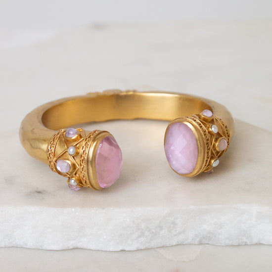 Gold cuff-style bracelet with two oval pink gemstones and small pearl-like accents, displayed on a white marble surface against a light neutral background.