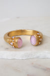 Gold cuff-style bracelet with two oval pink gemstones and small pearl-like accents, displayed on a white marble surface against a light neutral background.