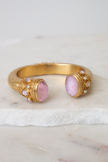 Gold cuff-style bracelet with two oval pink gemstones and small pearl-like accents, displayed on a white marble surface against a light neutral background.