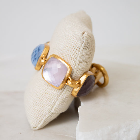 A gold-tone bracelet with glass stone details displayed on a beige fabric pillow over a white stone slab.
