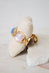A gold-tone bracelet with glass stone details displayed on a beige fabric pillow over a white stone slab.