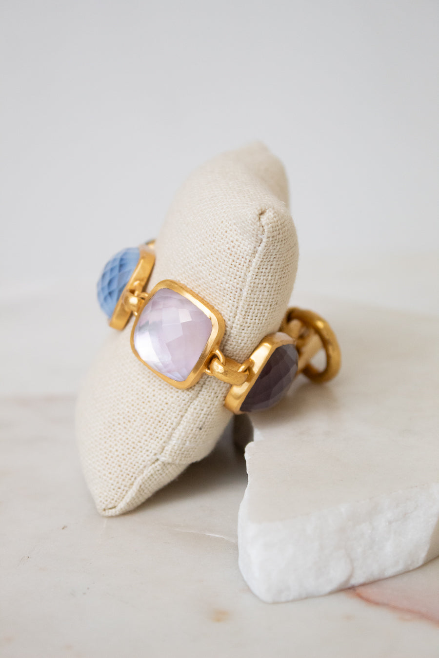A gold-tone bracelet with glass stone details displayed on a beige fabric pillow over a white stone slab.