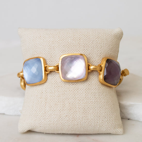A gold-tone cuff bracelet with three large square cabochon glass stones in light blue, milky white, and dark purple, displayed on a beige fabric jewelry pillow over a white marble surface.